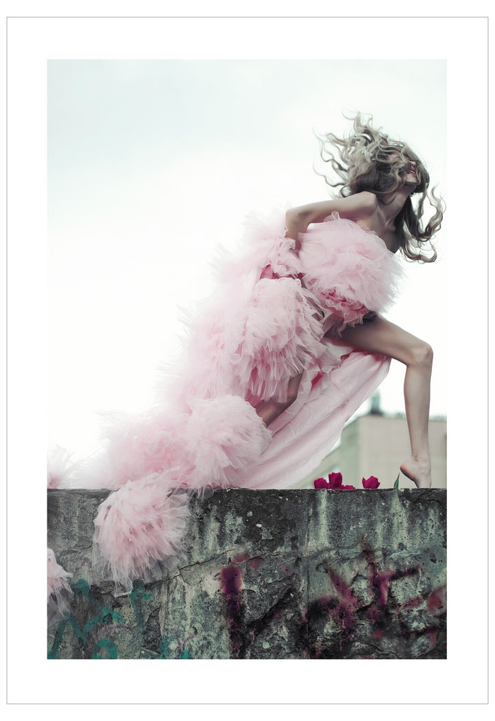 Woman in a pink dress with feathered skirt standing on a wall with graffiti.