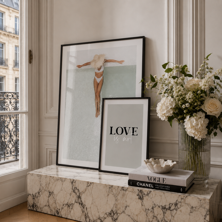 Two framed art prints displayed on a marble console table in a styled interior setting. One print features a minimalist fashion illustration of a woman, and the other features the text 'LOVE is art' in black lettering.