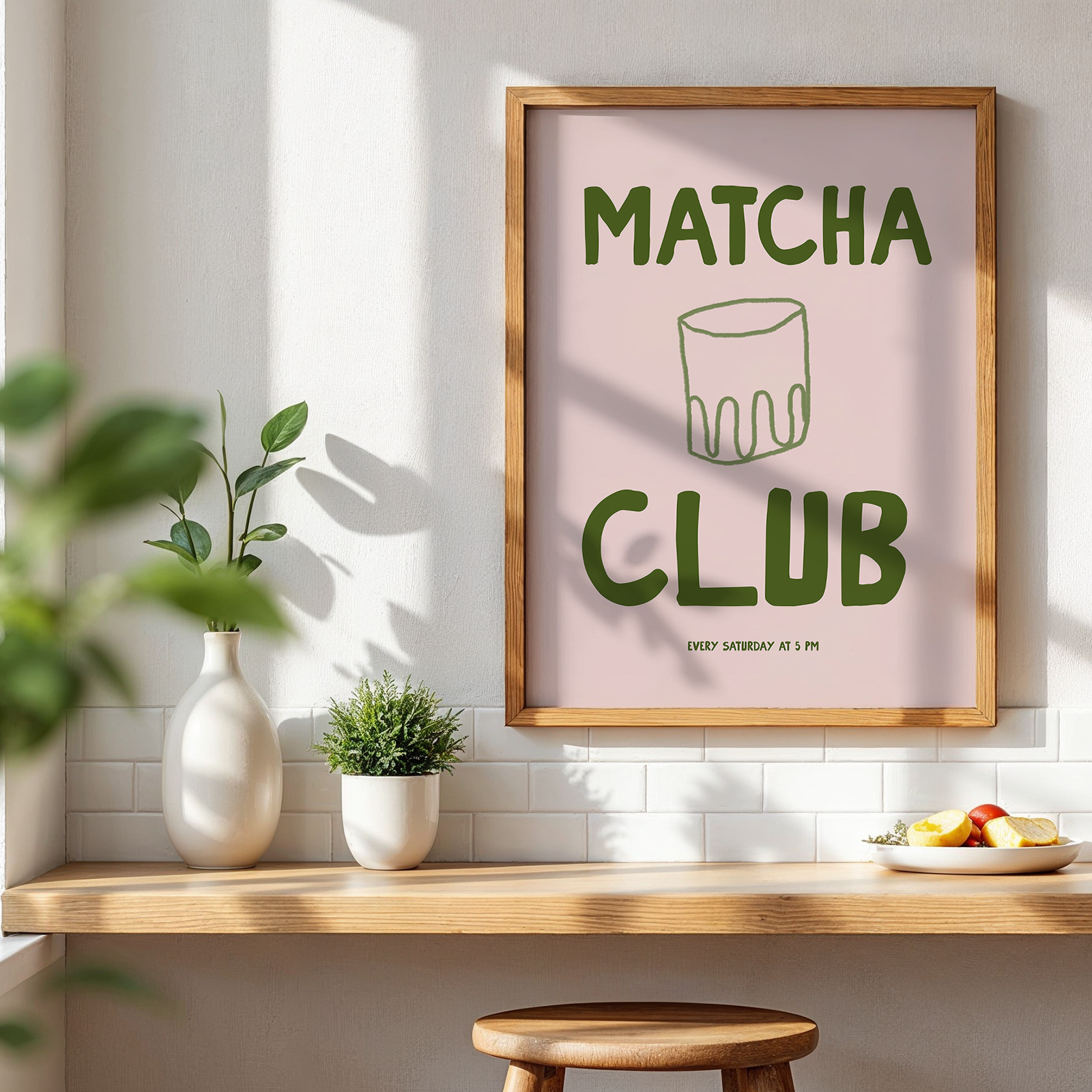 Framed poster with 'Matcha Club' text on a wall in a bright room with plants and a table.