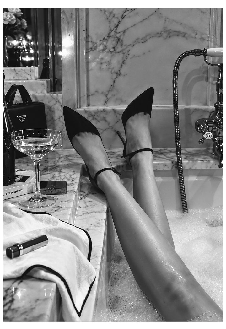 Black pointed-toe high heels with ankle straps worn by a person reclining in a bubble bath, with marble walls and a cocktail glass nearby.