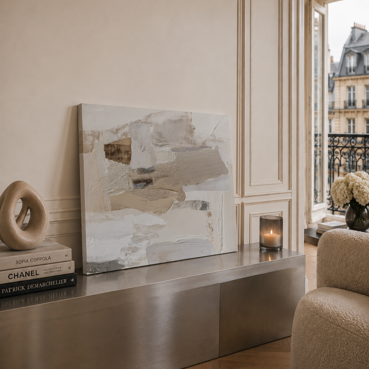 An abstract canvas painting with textured brushstrokes in beige, gray, and white tones, displayed on a metallic console table in a stylishly decorated room.
