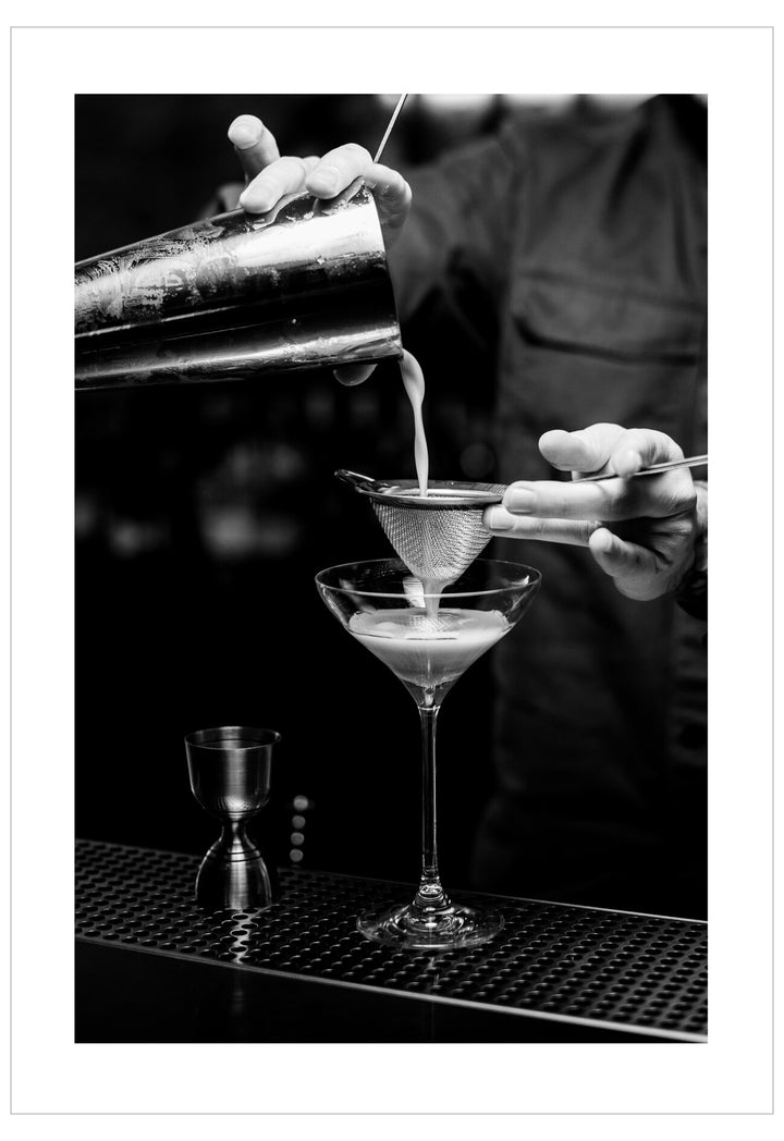 Black and white photo of a bartender pouring a cocktail into a glass.