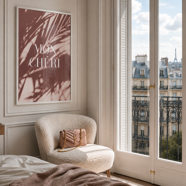 A framed wall art print featuring a dusty rose and brown palm shadow pattern with the text 'MGA CHERI' in white, displayed in a bright bedroom corner next to a window with a view of Paris rooftops. A cream boucle armchair with a pink handbag sits below the artwork.