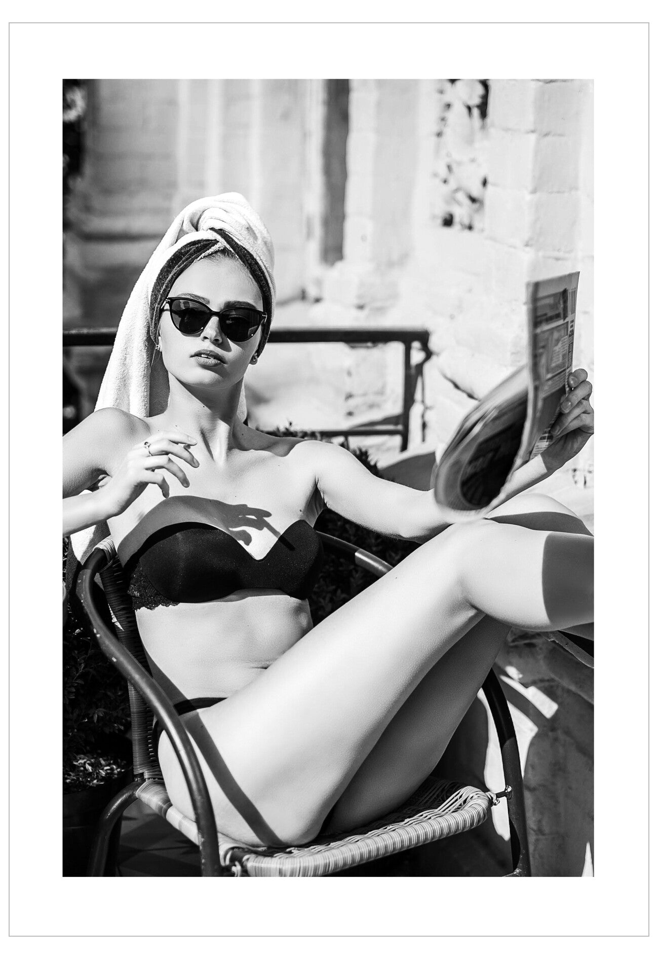 Woman in a black bikini and sunglasses lounging on a chair with a towel on her head, holding a newspaper.