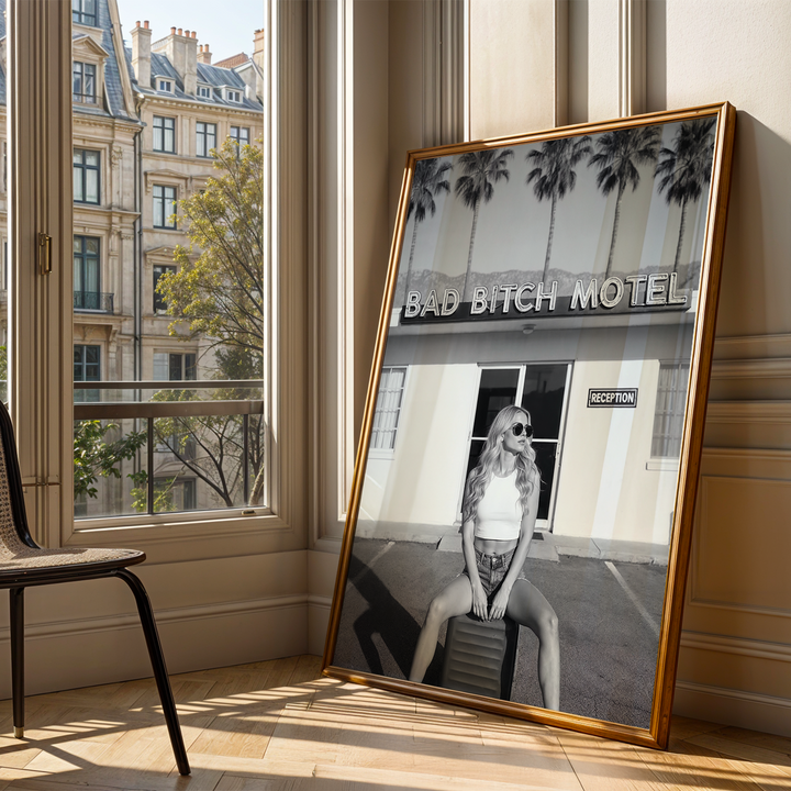 Framed artwork of a woman sitting on a stool with text 'BAD BITCH MOTEL' displayed in a room with large windows.