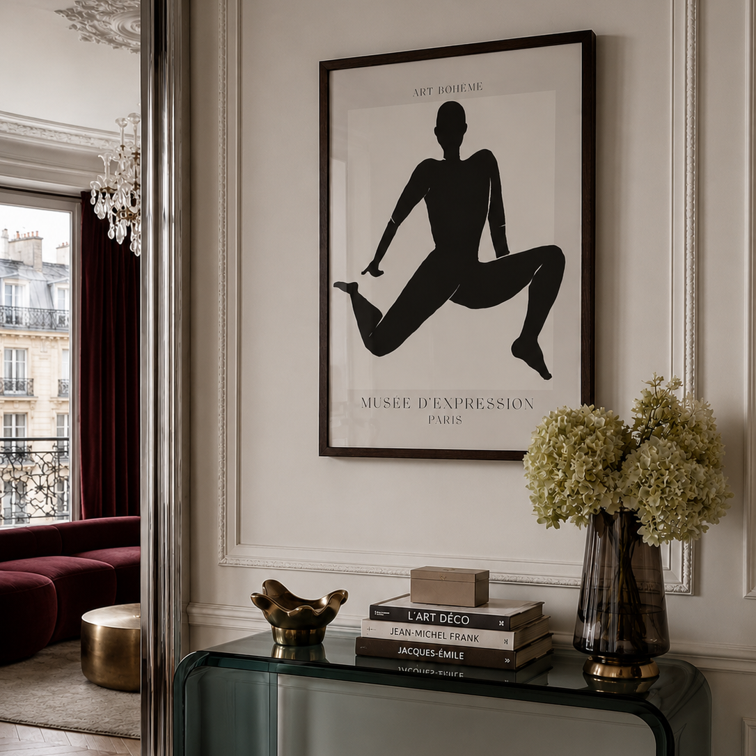 A framed black and white art print featuring a dancer silhouette and the text 'MUSÉE D'EXPRESSION PARIS' hanging on a white paneled wall in an elegant interior. Below the frame, a glass console table holds books, a small gold sculpture, and a vase of pale green hydrangeas.
