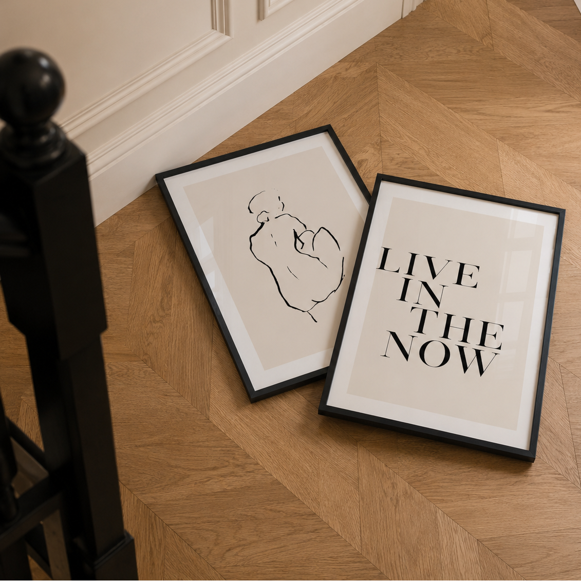 Two framed prints on a wooden floor, one with a black and white illustration and the other with 'Live in the Now' text.
