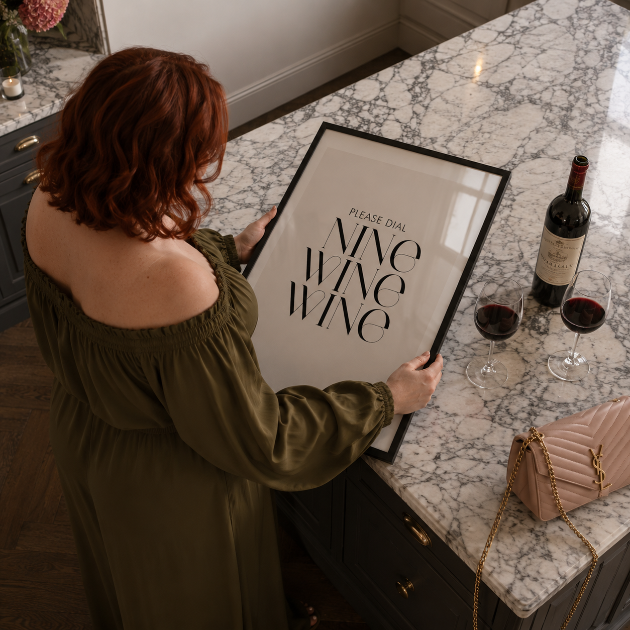 Woman holding a framed sign with 'Please dial NINE WINE WINE' on a marble countertop with wine glasses and a bottle.