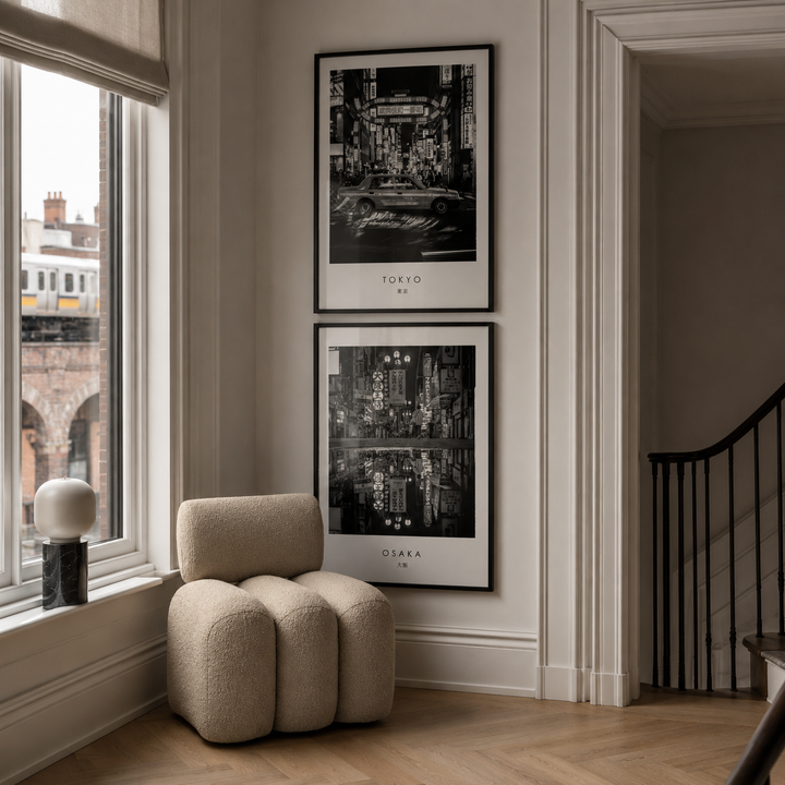 A styled interior corner with two framed black-and-white cityscape prints hanging on a beige wall above a rounded cream boucle armchair. The prints read Tokyo, Japan and Osaka, Japan.