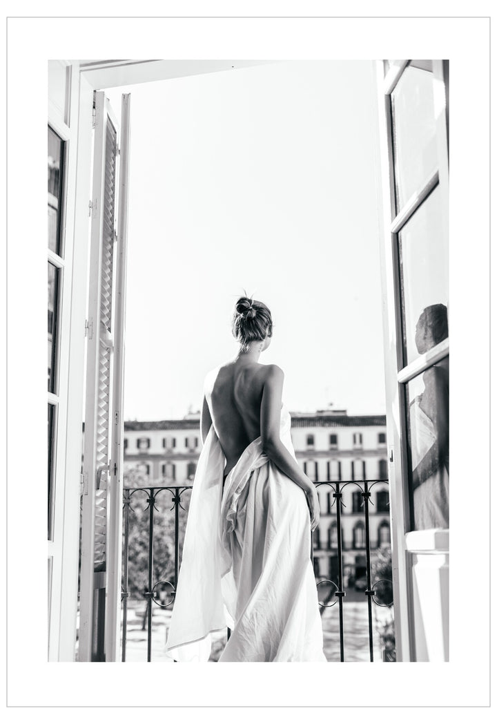 Woman in a flowing dress standing by an open window with a cityscape view.