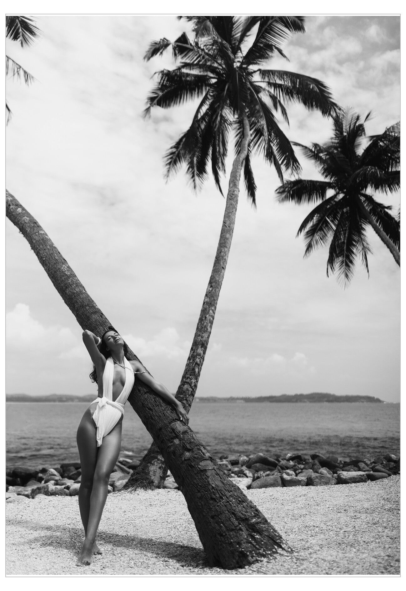 Person in a swimsuit leaning against a palm tree on a beach with ocean and sky in the background