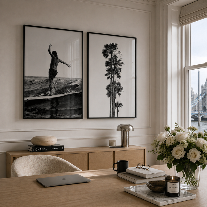 Two framed black-and-white wall art prints hang above a wooden console in a bright, modern room. The left print shows a surfer on a wave, and the right print shows tall palm trees.