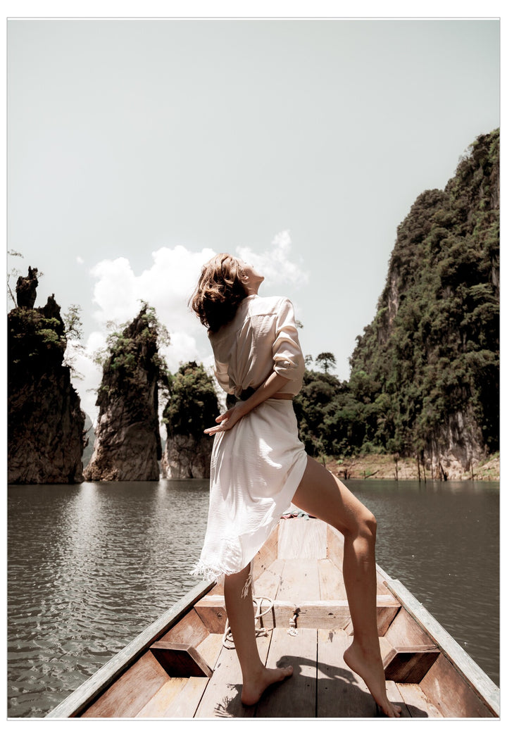 Woman in a white dress standing on a wooden boat with a scenic background of water and cliffs.