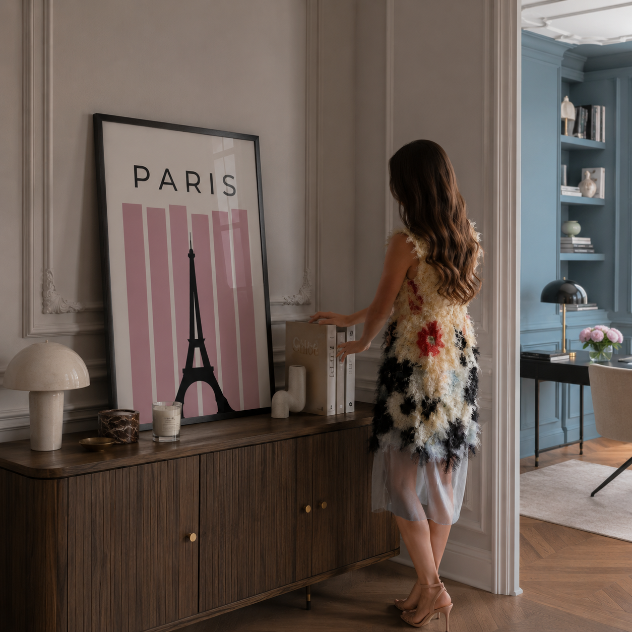 A woman stands in a doorway wearing a sleeveless Chanel Haute Couture cream dress with black and white feather-like appliqué and a small red accent, while looking at a large framed Paris poster inspired by Emily in Paris leaning on a wooden console table in a styled interior.