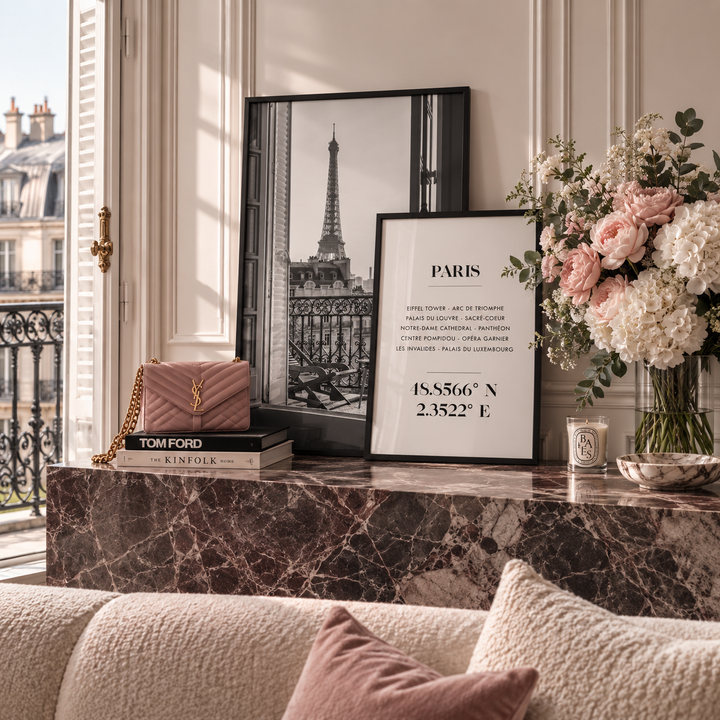 A styled interior scene with framed wall art leaning on a marble console table. The most prominent sellable subject appears to be the Paris-themed framed print/poster in the center-right, showing a black-and-white Eiffel Tower image and Paris coordinates, with additional decor including flowers, a pink quilted handbag, books, and a candle in a nearby room.