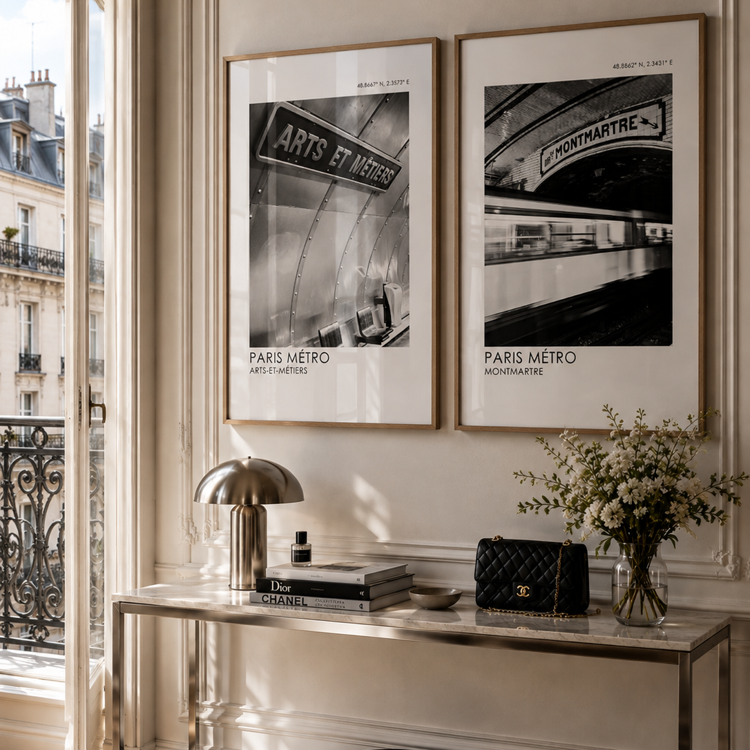 Two framed black and white Paris Metro art prints hang on a beige wall above a console table in a sunlit room. The scene includes a metallic lamp, books, a black quilted handbag, and a vase of flowers.
