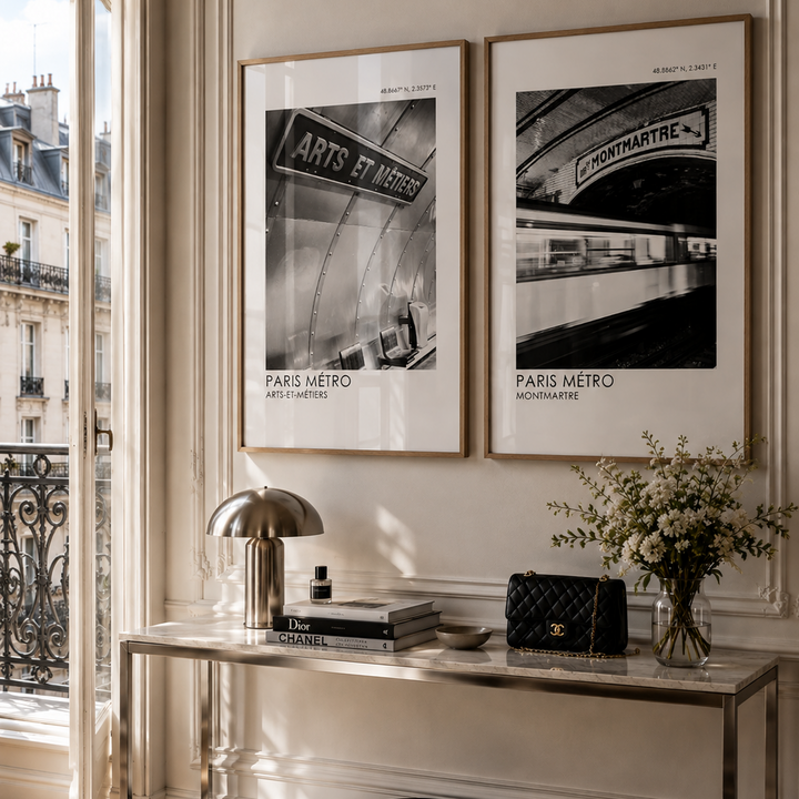 Two framed black and white Paris Metro art prints hang on a beige wall above a console table in a sunlit room. The scene includes a metallic lamp, books, a black quilted handbag, and a vase of flowers.