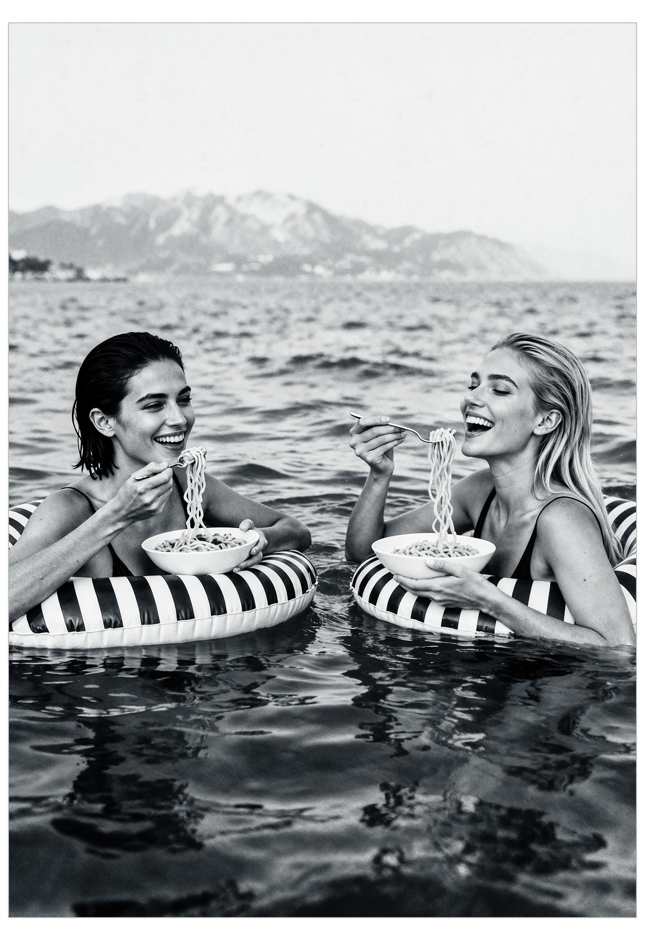 Two women on striped inflatable rings in water with mountains in the background