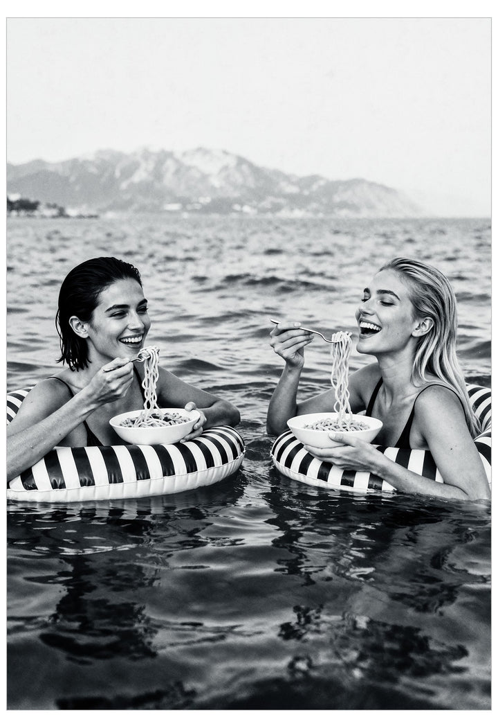 Two women on striped inflatable rings in water with mountains in the background