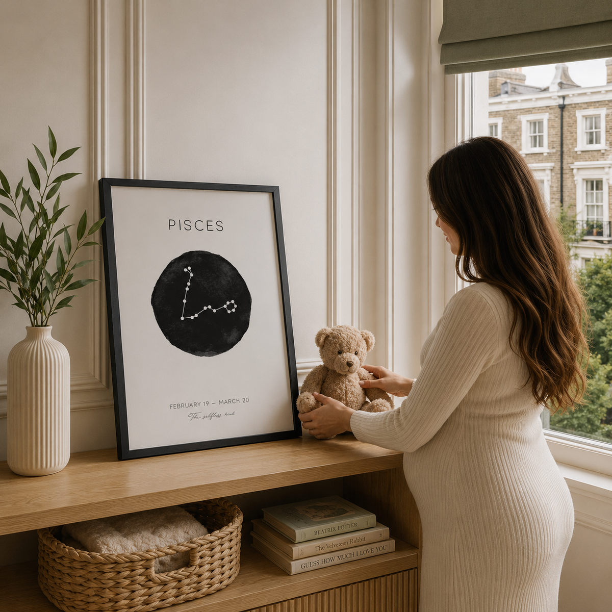 A woman in a ribbed cream dress stands by a window beside a framed Pisces zodiac art print on a wooden shelf, holding a teddy bear. The room has warm neutral tones, a vase with greenery, and a window view.