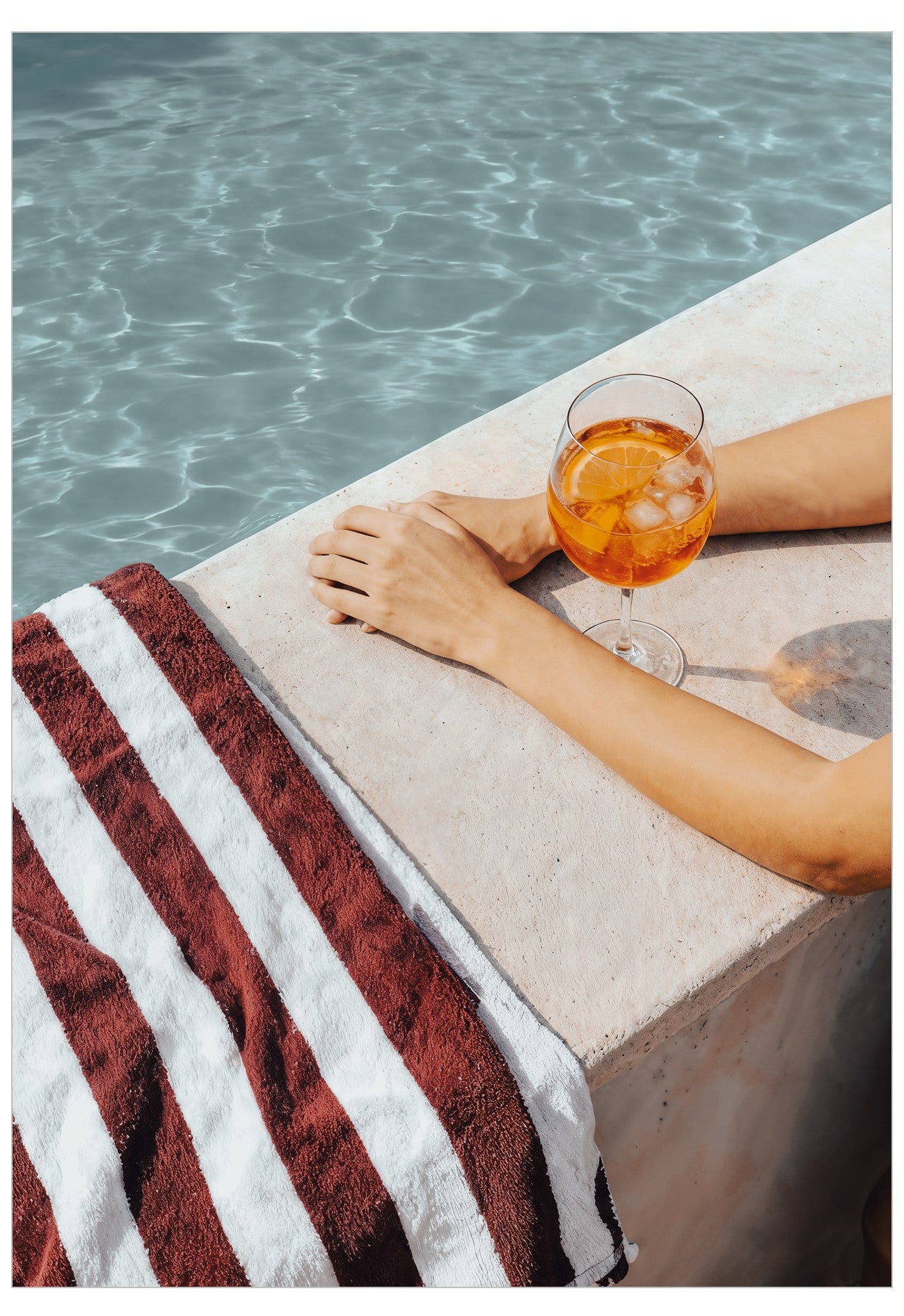 Person holding a glass of orange aperol spritz cocktail by a poolside with a striped towel nearby.