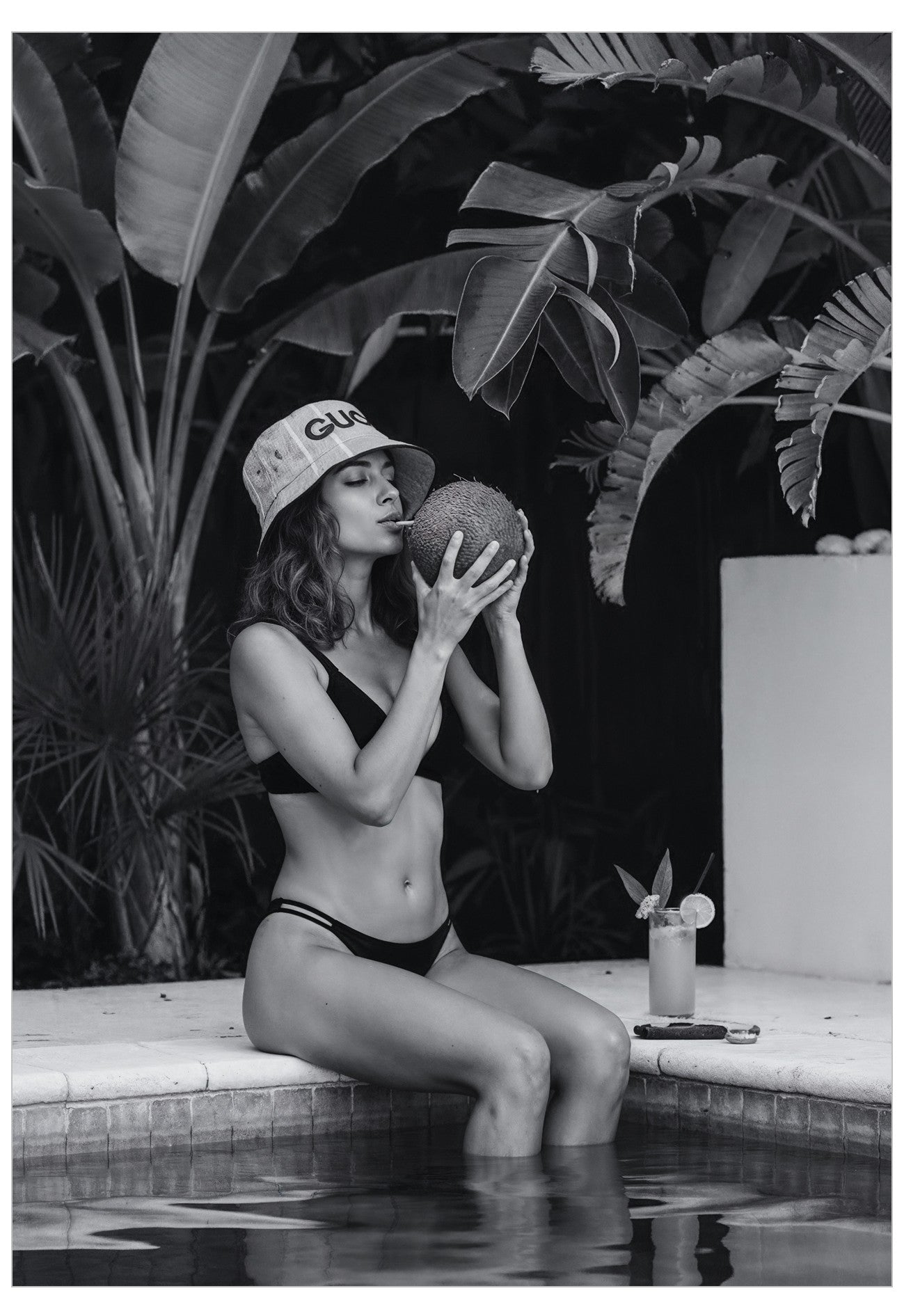 Person sitting by a pool holding a coconut wearing a gucci bucket hat, surrounded by tropical plants.