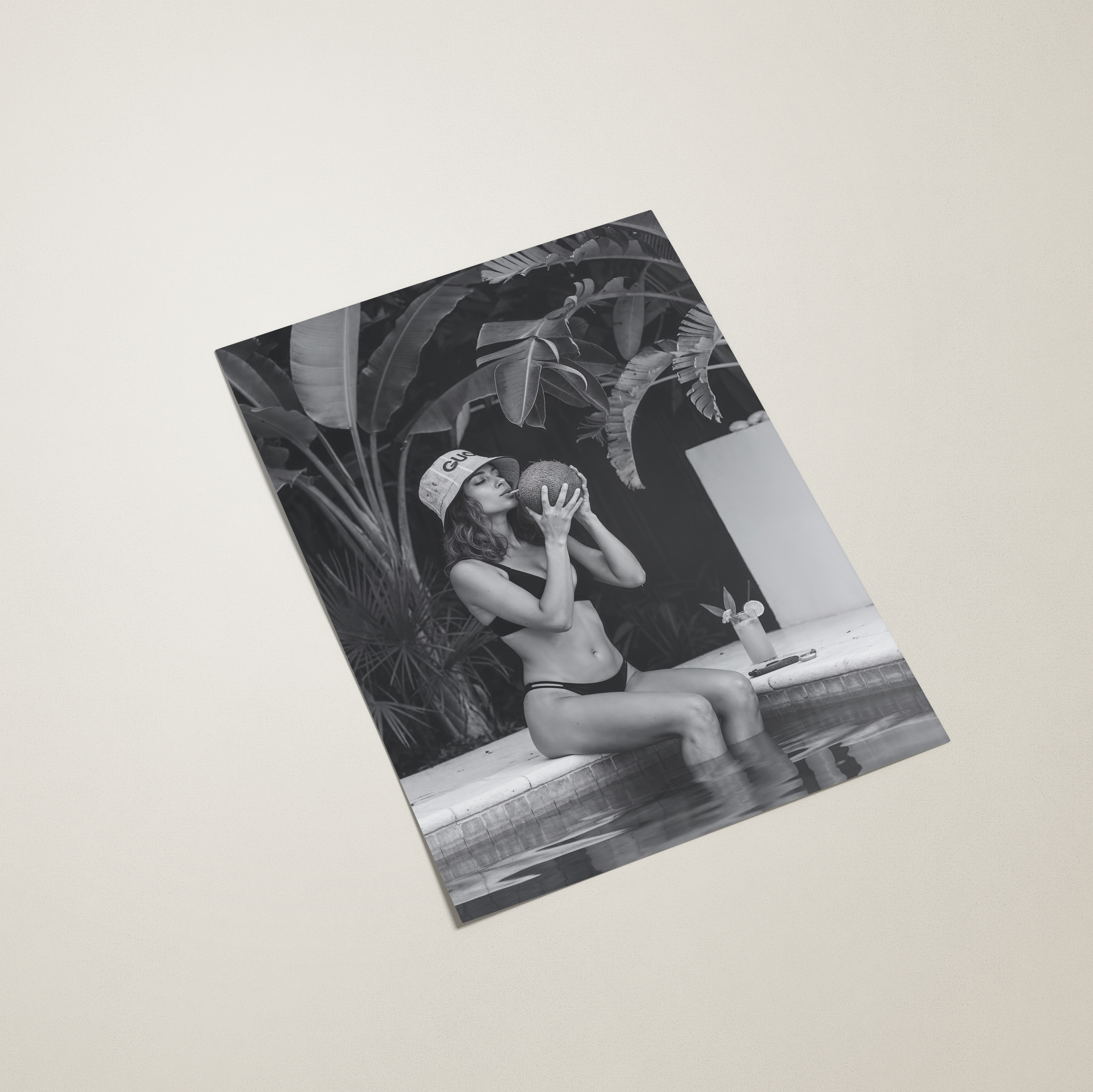 Black and white photo of a woman sitting by a pool wearing a gucci bucket hat with plants and a building in the background.