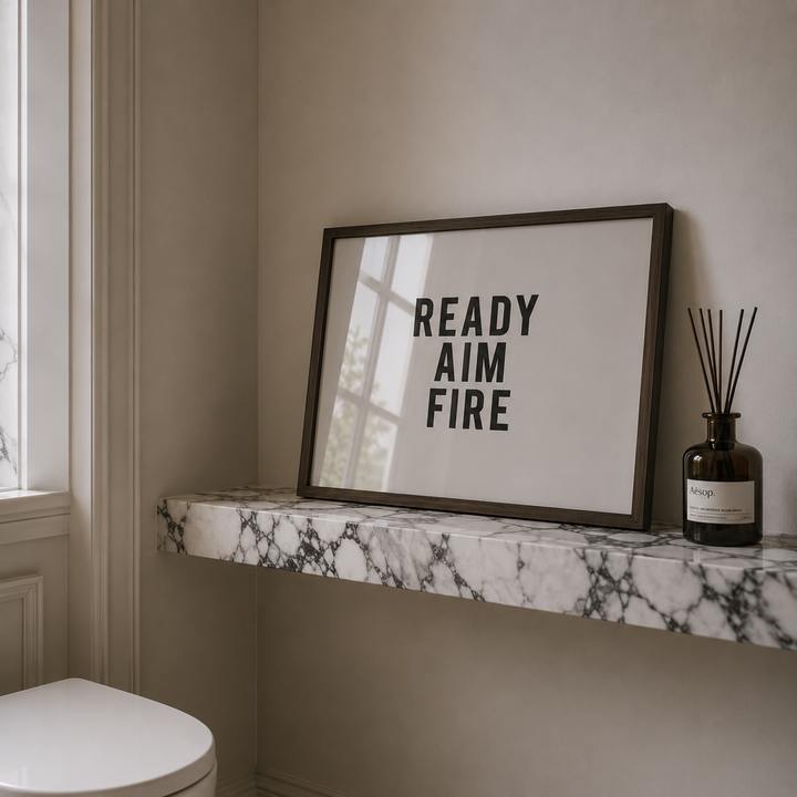 Framed poster with 'READY AIM FIRE' text on a marble shelf in a bathroom.