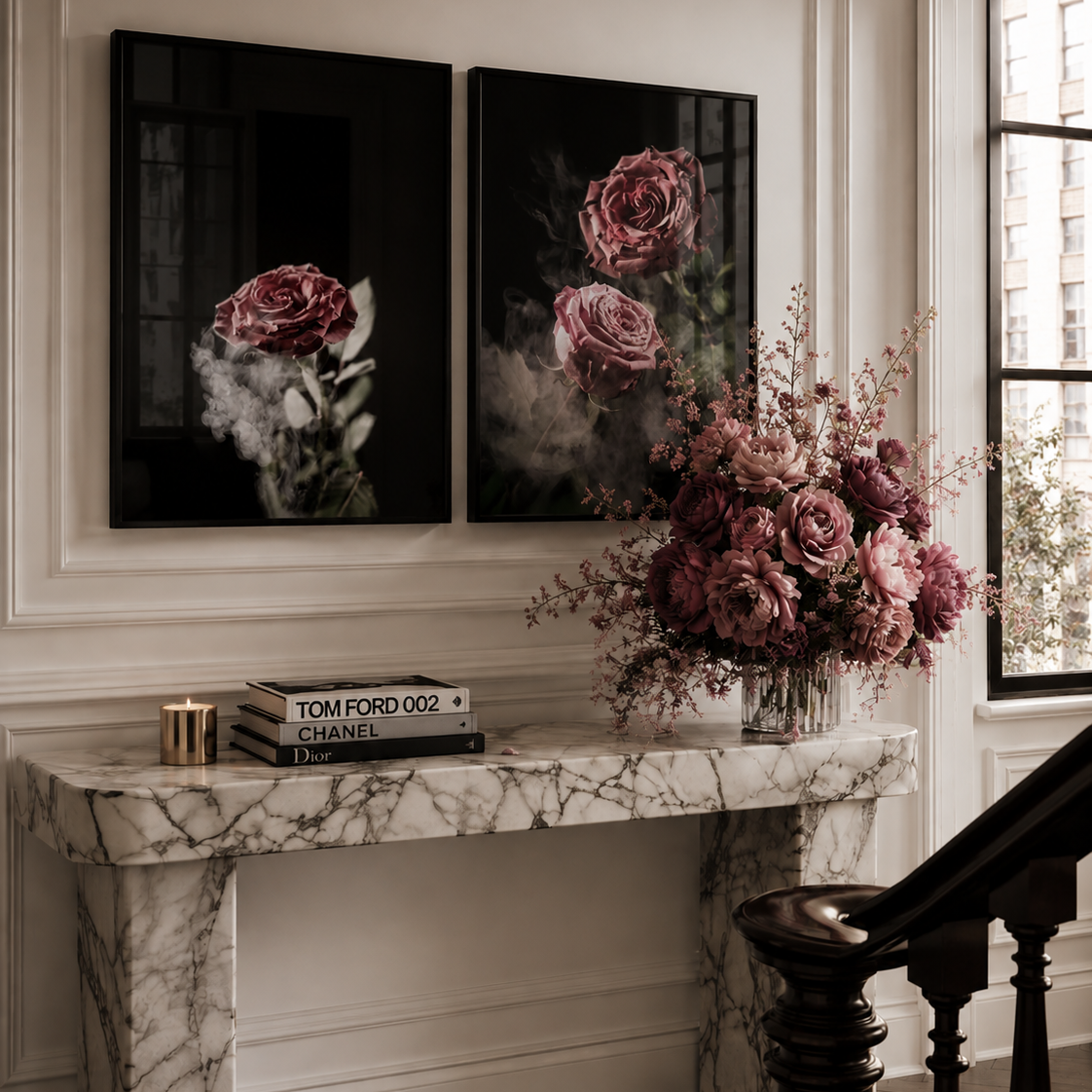 A styled interior scene showing two vertical black canvas prints with pink rose imagery mounted on a cream wall above a marble console table. A large pink floral arrangement sits on the table, with books and a candle nearby, and a dark wooden staircase railing in the foreground.