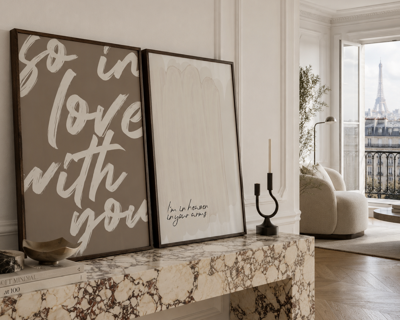 Two framed art prints leaning against a white wall on a marble mantel in a styled living room. The left print has dark brown and white script text reading 'so in love with you', and the right print has a soft white brushstroke background with small black handwritten text.