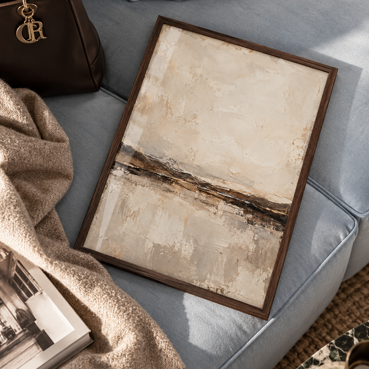 A framed abstract landscape painting with beige, cream, and brown tones, styled on a light blue sofa next to a brown leather handbag and a beige knit throw blanket.
