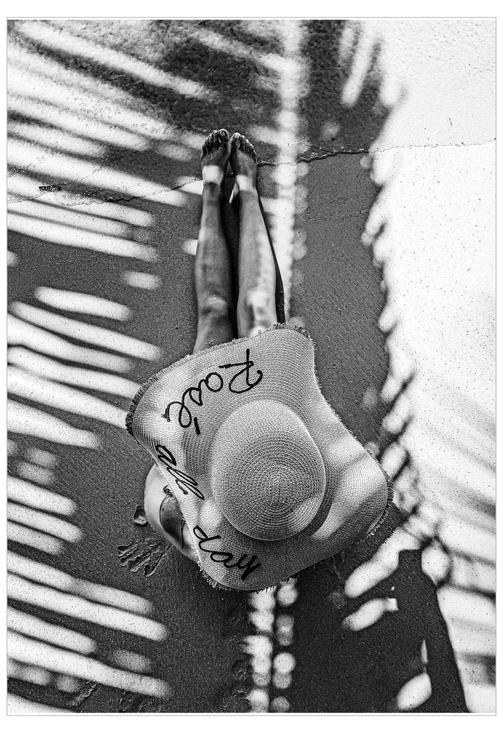Black and white photo of a person wearing a wide-brimmed hat with text, standing against a palm leaf background.