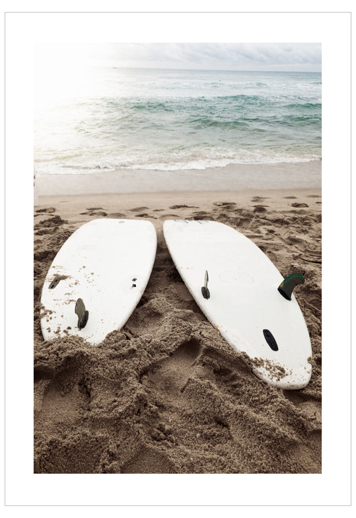 Two white surfboards lying on a sandy beach with ocean waves in the background.