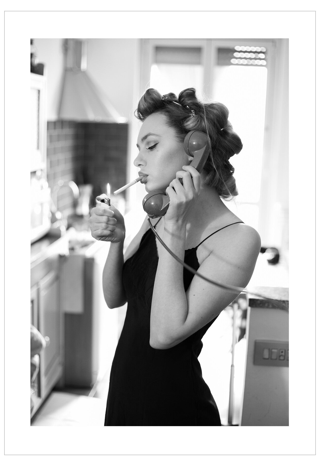 Woman in a black dress talking on a vintage phone and smoking a cigarette in a kitchen.