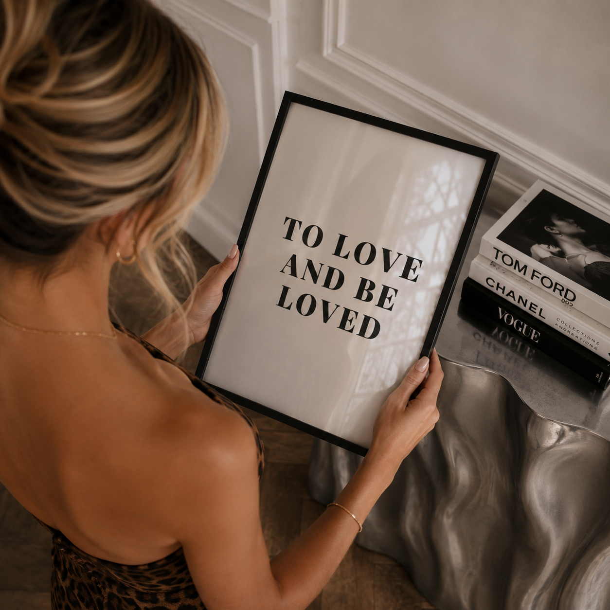 Woman holding a framed quote 'TO LOVE AND BE LOVED' in a stylish room.
