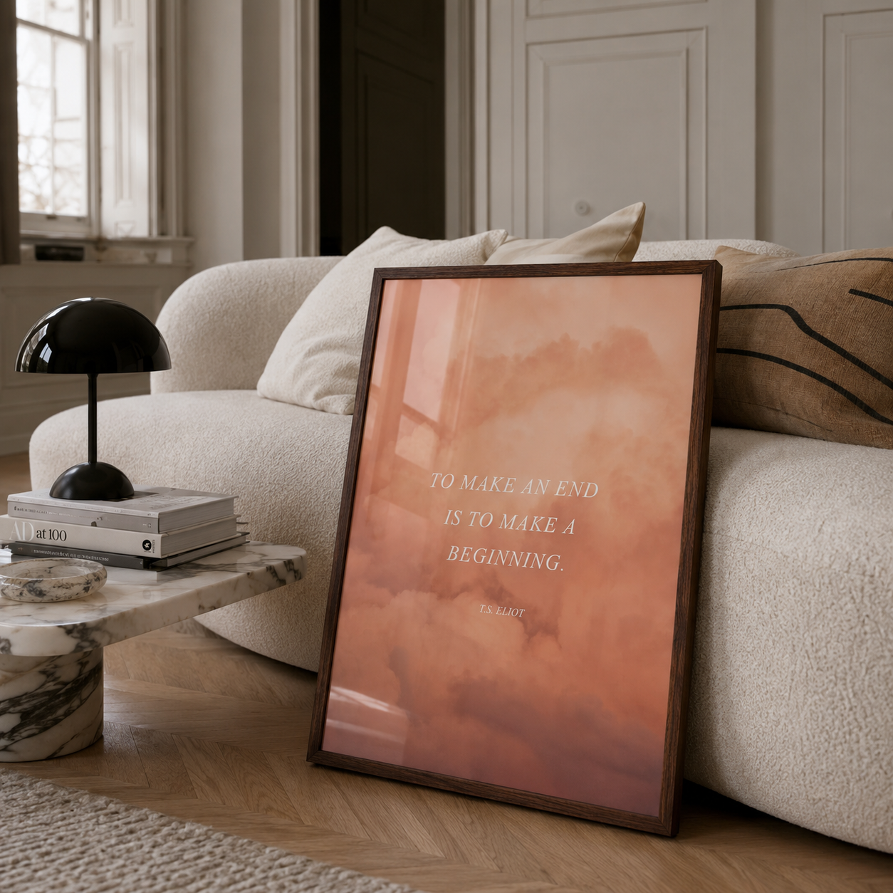 A framed art print with a warm peach and orange abstract design and the quote 'TO MAKE AN END IS TO MAKE A BEGINNING.' leaning against a beige boucle sofa in a modern living room.