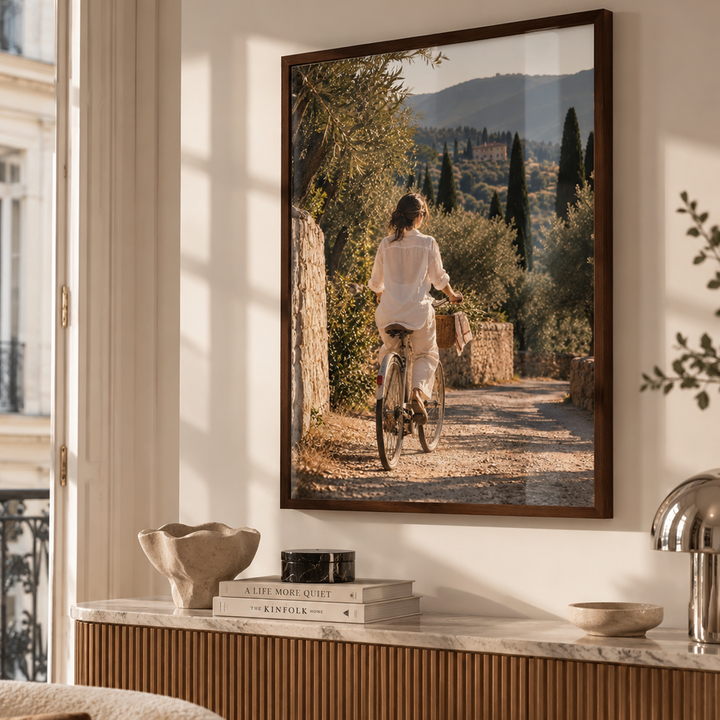A framed vertical art print depicting a woman riding a bicycle down a sunlit stone path with olive trees and hills in the background, displayed on a wall above a console table in a modern, warm-toned living room.