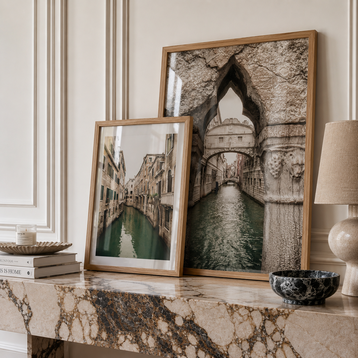 Two framed art prints depicting scenic Venice canals and stone arches, styled on a marble mantelpiece in a home interior setting.