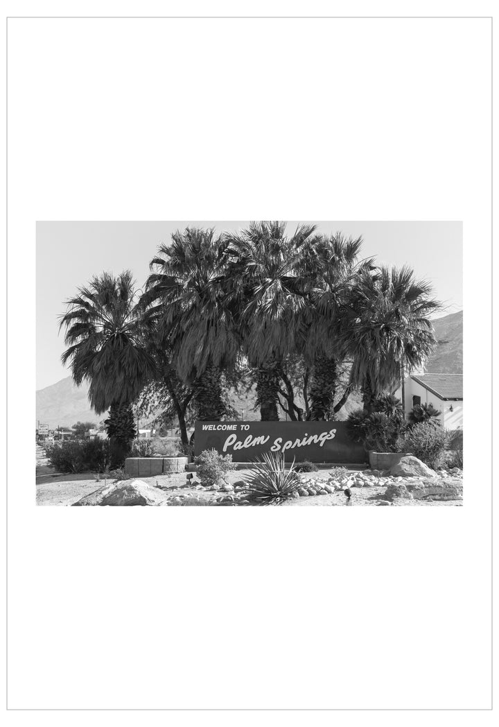 Black and white photo of a 'Welcome to Palm Springs' sign with palm trees.