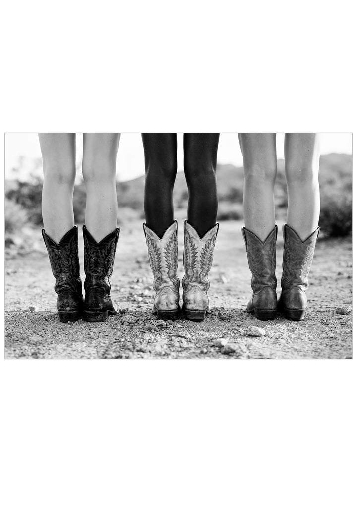 A black and white photograph showing the lower legs of three people standing outdoors on a gravelly ground, each wearing a pair of cowboy boots. The center figure wears patterned western boots, flanked by two figures in darker cowboy boots.