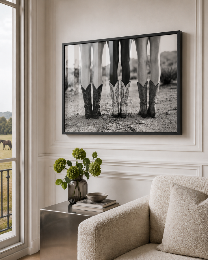 A framed black and white photographic print of three pairs of cowboy boots hanging on a cream-colored wall in a luxury styled living room setting.