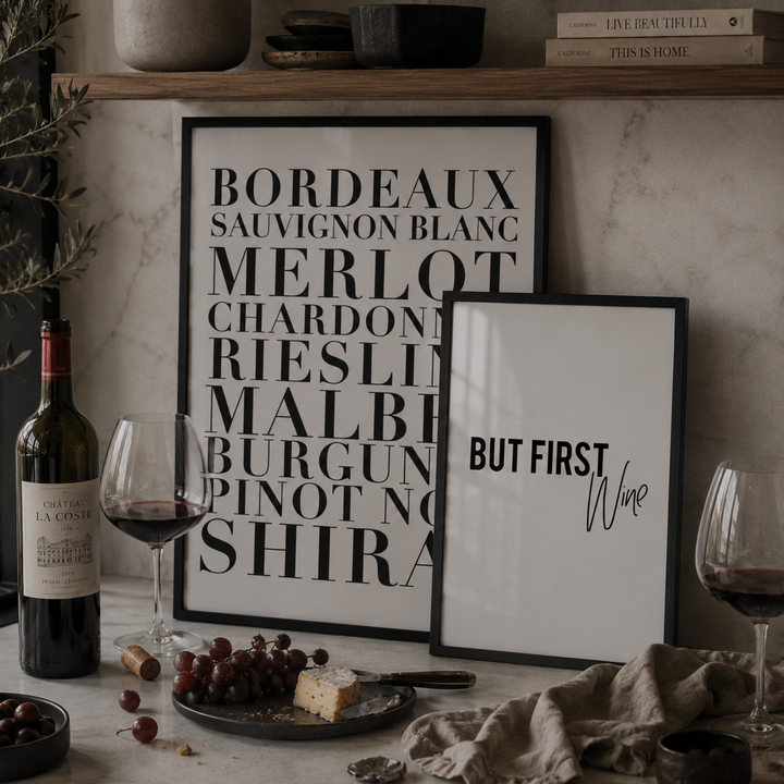 Wine bottle, glasses, and grapes on a table with framed art in the background.
