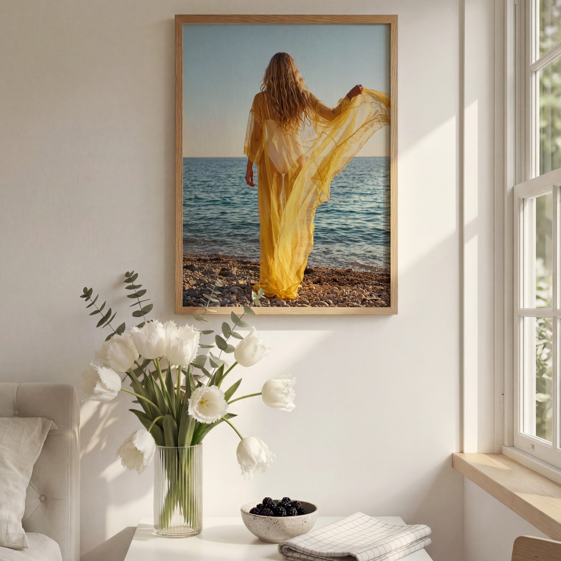 Framed artwork of a woman in a yellow dress on a beach, above a vase with white flowers and a bowl of fruit in a room with a window.