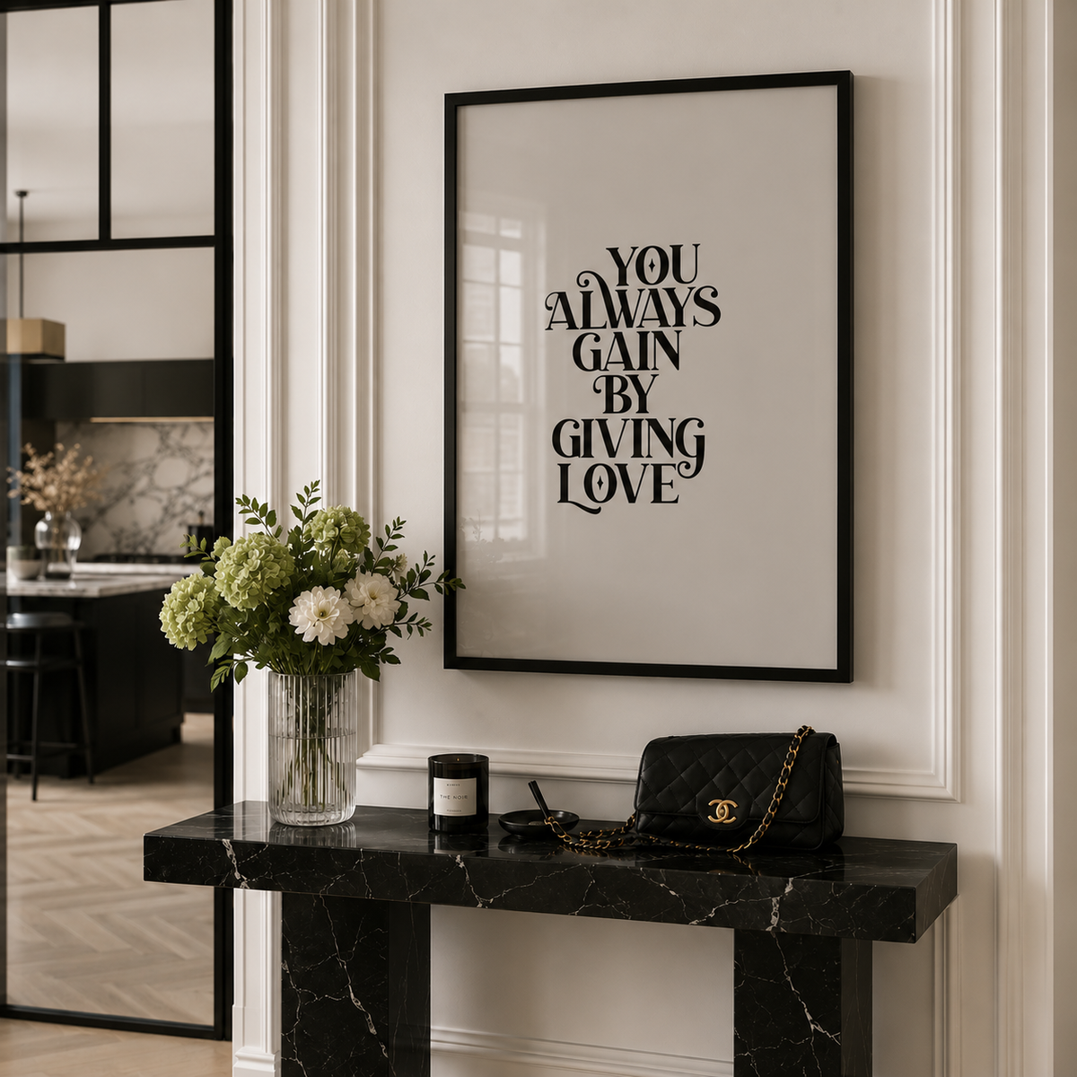 A framed typographic wall art print with the phrase 'YOU ALWAYS GAIN BY GIVING LOVE' in black lettering on a light beige background, hanging above a black marble console table in a styled interior. A vase of flowers, a candle, and a quilted black handbag are placed on the table.