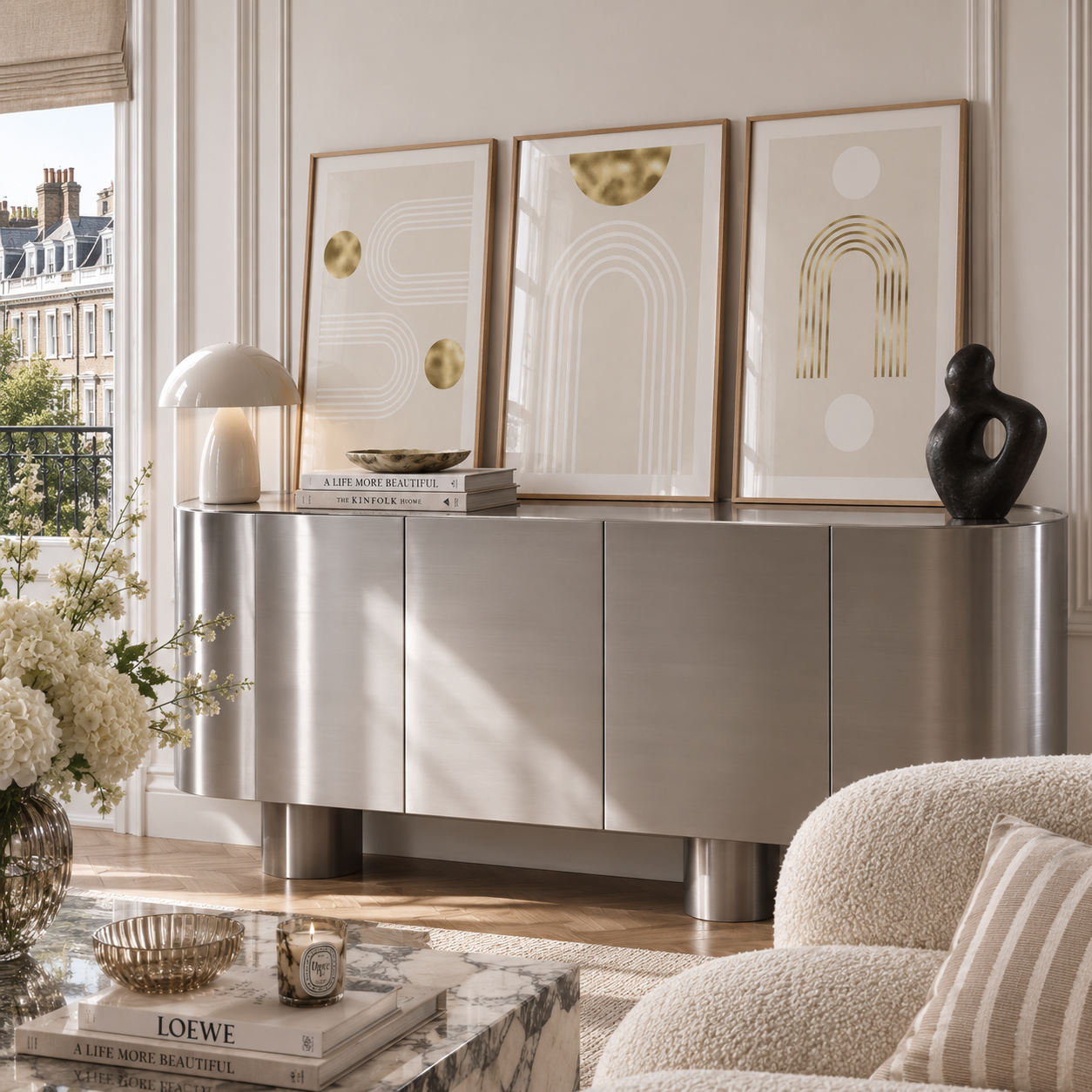 A modern silver sideboard or console cabinet in a bright living room, styled with three framed abstract prints, a white mushroom lamp, books, a black sculptural vase, and a floral arrangement.