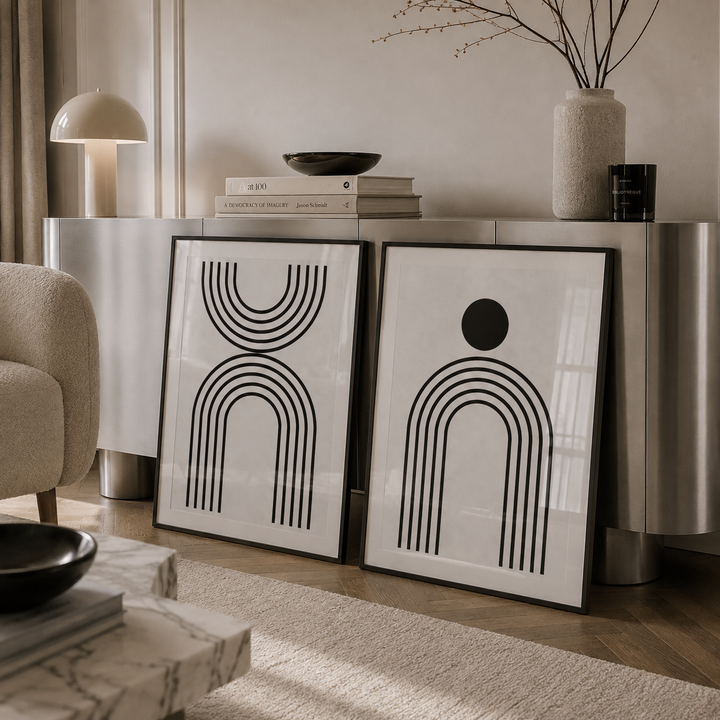 Two framed abstract black line art prints leaning against a metallic console table in a modern living room setting.