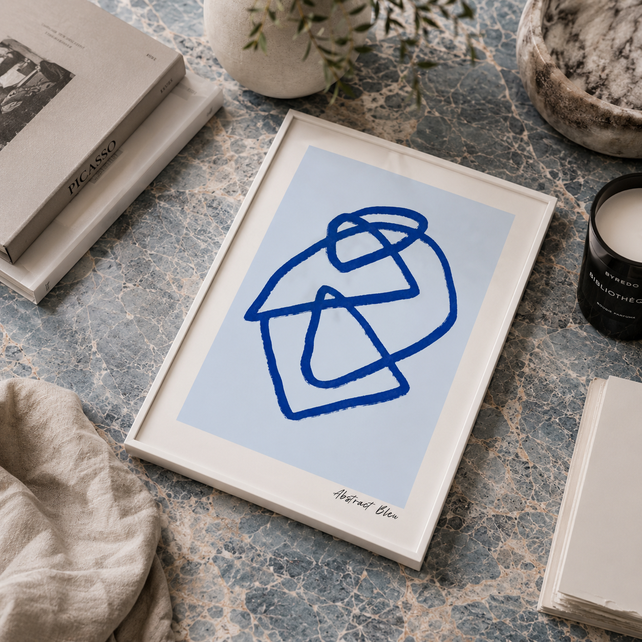 A framed abstract art print featuring a blue sketch on a light blue background, styled on a blue-gray marble surface with books, a candle, and a linen cloth.