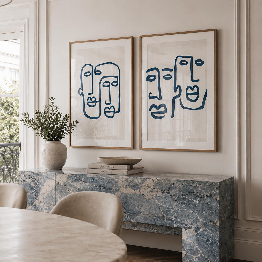 Two framed abstract face line art prints in navy blue on a white background, displayed side-by-side on a blue marble console table in a dining room setting.