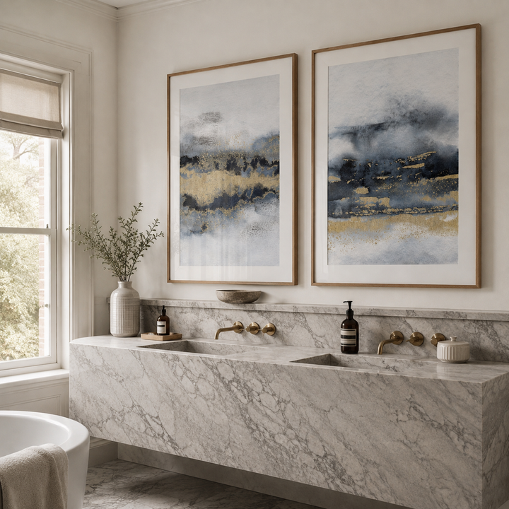 Two framed abstract landscape art prints with blue, gray, and gold tones hang above a double-sink marble bathroom counter in a bright, modern bathroom.