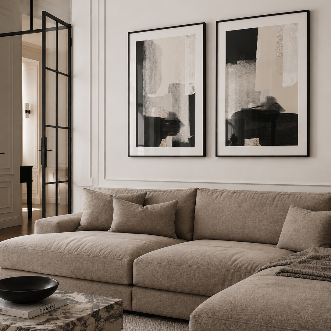 A modern beige sectional sofa in a styled living room, featuring two large black and beige abstract framed prints on the white wall above it.